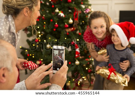 A father is taking a picture of his son and daughter, standing in front on the Christmas Tree.