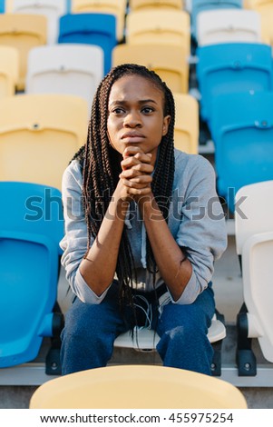 Worried african american young woman sitting waiting in rows of empty seats at stadium with her chin resting on hands and a glum expression