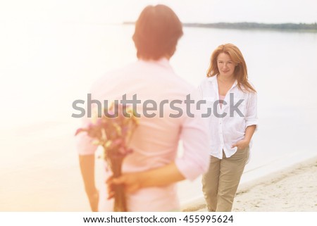 Toned picture of romantic couple walking on sandy beach. Handsome man hiding bouquet of flowers for his wife. Beautiful red-haired woman reaching to him with hands in pockets.