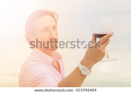 Toned picture of sommelier analyzing glass of red wine isolated on blue sky background. Black-haired mature man happy looking at glass.