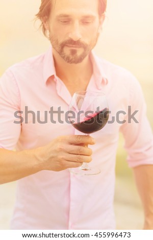 Sommelier tasting expensive old red wine. Toned picture of bearded man in pink shirt looking at glass with delicious red wine.