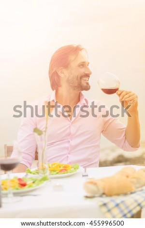 Toned picture of handsome man drinking red wine while sitting in restaurant or cafe by sea. Handsome bearded man in pink shirt relaxing and enjoying bunny day.
