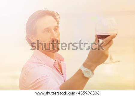 Toned picture of sommelier analyzing a glass of red wine isolated on blue sky background. Black-haired mature man happy looking at glass.