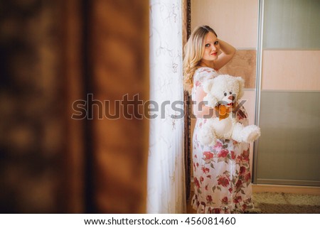 Pretty pregnant woman lying on the bed and touching her stomach. Woman stay near window. with teddy bear toy. Make up.