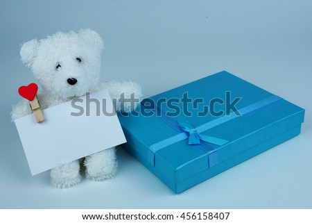 Close up of blue giftbox with blue ribbin and white cutty teddy bear holding white card on white background.