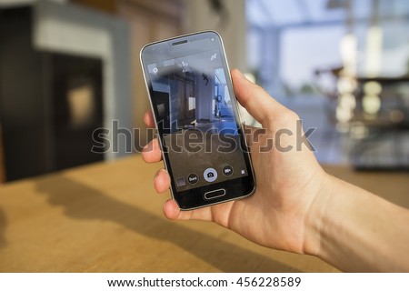 A hand holding a mobile phone which displays the camera app on the touch screen. The image is taken in the living room of a home.