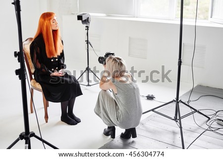 Girl the photographer takes pictures of model in black sitting on a chair on white background in Studio
