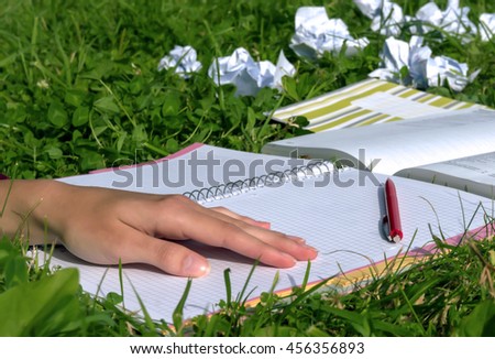 girl in a burgundy sweater is put her hands on the book, which lies on the green grass, notebook empty, a creative crisis, is located next to the red pen and book