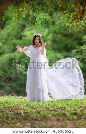 beautiful young woman in white dress with flower on her hair playing violin outdoor shot. Portrait of beautiful blond young woman standing on green tree background