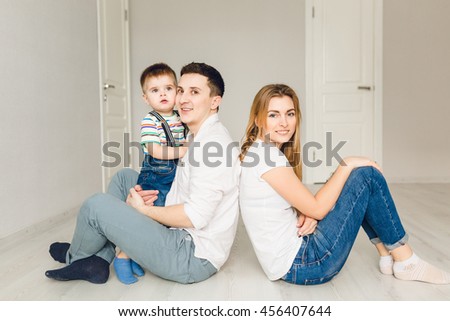 Family picture of two young parents playing with their boy child. They sit on the floor and father holds his son. They wear white t-shirt and jeans.