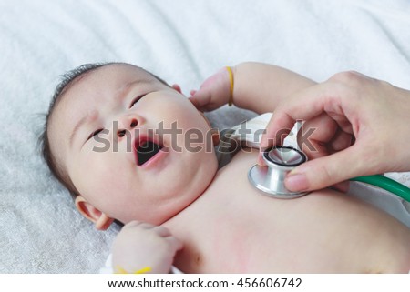 Professional pediatrician examining infant. Doctor using a stethoscope to listen to kid's chest checking heartbeat. Two months baby asian girl lying on sickbed in hospital.