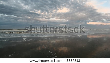 Sunset, waves on the seashore, pink and blue clouds reflect in wet broad sandy seashore.