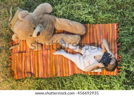 Young little girl photographer with old vintage film photo camera laying down on picnic blanket with teddy bear taking picture outdoor. Children's play. Art or creativity concept.