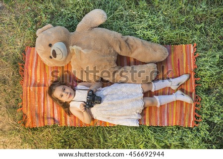 Young little girl photographer with old vintage film photo camera laying down on picnic blanket with teddy bear outdoor in a sunny summer day. Children's play. Art or creativity concept.