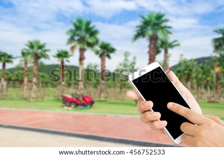 woman use mobile phone and blurred image of a man drive red mower on the field with palm trees , mountains , and blue sky for background
