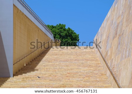 Wide staircase built from stones of yellowish colors in the historic city of Valletta, Malta, on a background of clear blue sky and green crown of tree.