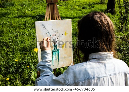 Young female artist painting flowers in bottle on canvas in summer field