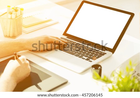 Closeup of male hands using laptop with blank screen and graphic tablet on office desktop with telephone, stationery items and plant. Toned image, Mock up