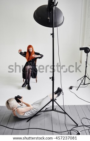 Girl photographer lies on the floor and shoots of fashion model posing on chair on white background in Studio