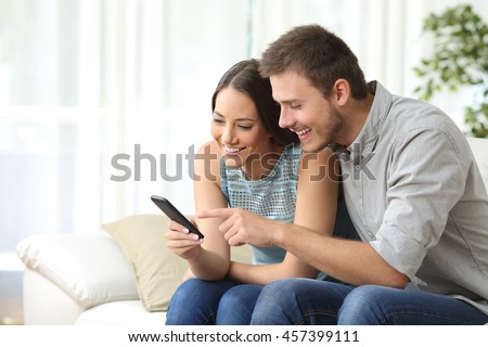 Relaxed couple or friends using a generic mobile phone together sitting on a sofa in the living room at home Royalty-Free Stock Photo #457399111