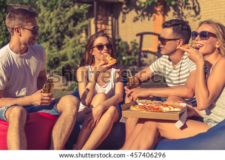 Young beautiful people in casual clothes and sun glasses are eating pizza, communicating and smiling, sitting on bean bag chairs while resting outdoors