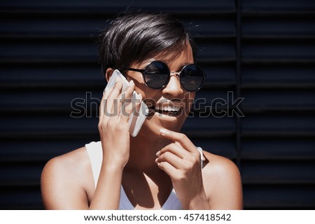 Outdoor portrait of fashionable African American woman in round shades talking on smart phone to her friend, discussing latest news, smiling, laughing, looking cheerful and excited, touching her chin