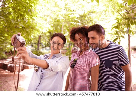 Young threesome looking and smiling at a camera to take a selfie in the dappled shade some trees wearing casual clothing