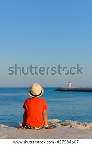 Back view of cute boy on the beach looking at ocean lighthouse on sunny outdoors background. Book cover concept design
