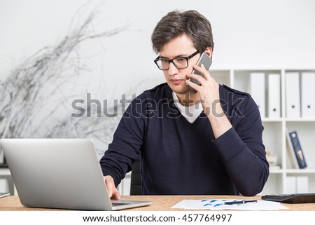 Young man in dark sweater and glasses working at his desk in office and talking on smartphone. Concept of business planning