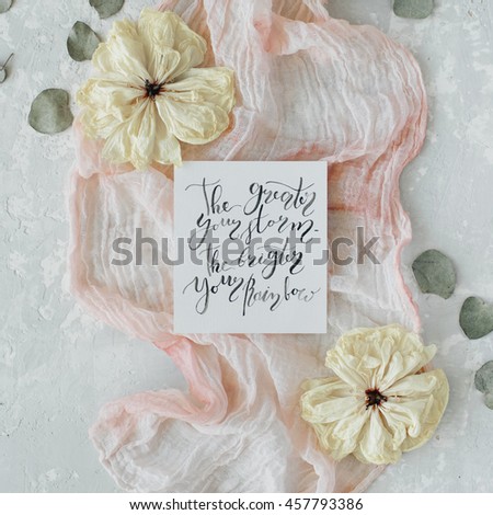 inspirational quote "the greater your storm the brighter your rainbow" written in calligraphy style on paper with dry white tulips, eucalyptus petals and pink textile on concrete background. Flat lay