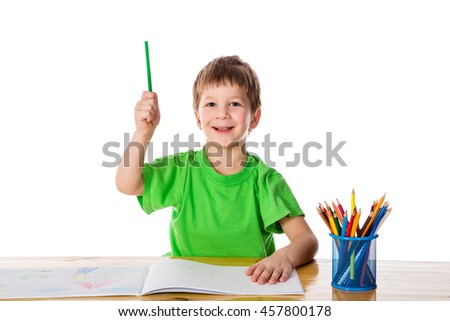 Creative smiling little boy at the table with pencils, pointing up, gesturing idea, isolated on white