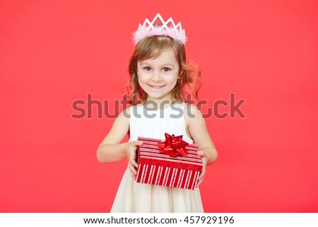 Little girl holding a gift box over red background. Blond hair little kid princess on bright vivid color wall