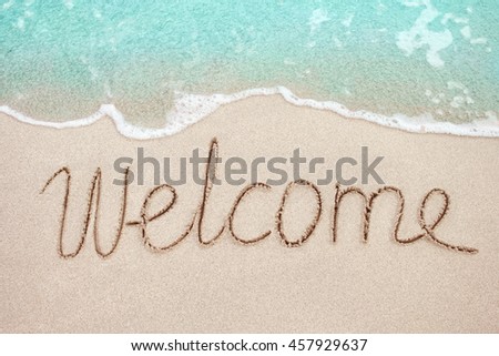 Word Welcome handwritten on sandy beach with soft wave of blue ocean on background.