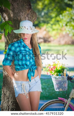 Portrait of female model posing by leaning against the tree in a park. Bicycle with basket full of flowers in the background. Summer, lifestyle and fashion concepts. Space for copy.
