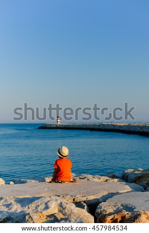 Back view of cute boy on the beach looking at ocean lighthouse on sunny outdoors background. book cover concept design
