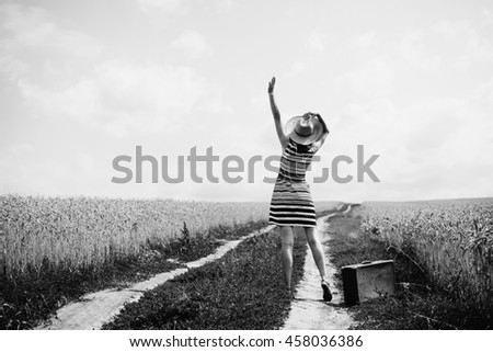 Black and white photography of happy young woman carrying suitcase waving to the sun. Lady with straw hat traveling along the road, rural outdoors background