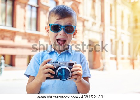 Small boy in sunglasses is making excited face while holding a photo camera and trying to take a shot during walk in historical part of the city