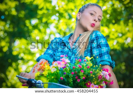 Portrait of pretty young female with pursed lips on bicycle with basket full of colorful flowers. Summer, nature, fashion and recreation concepts.  