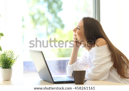 Pensive girl with a laptop daydreaming and looking through a window in a house with a green background outdoors in a beautiful day Royalty-Free Stock Photo #458261161