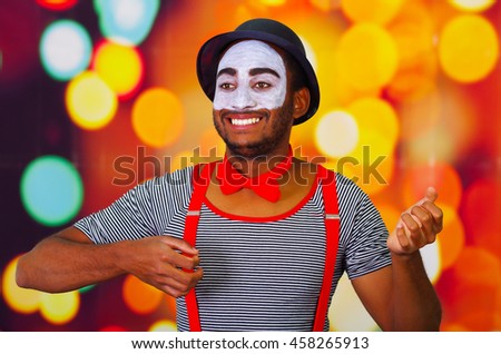 Pantomime man wearing facial paint posing for camera, using hands interacting body language, blurry lights background