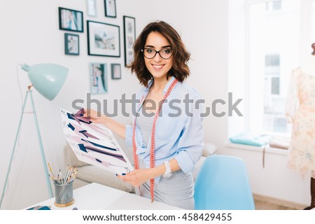 A brunette girl in a gray dress and blue shirt is standing near the table in a workshop studio. She has some samples and sketches in her hands. She is smiling to the camera.
