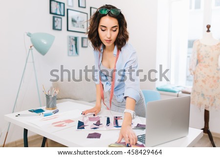 A brunette girl in a gray dress and blue shirt is standing near the table in a workshop studio. She has a lot of creative stuff  on the table. She is looking for some material samples.