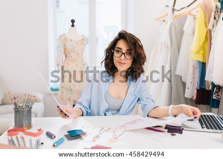 A brunette girl is sitting at the table in the workshop. She holds sketches in both hands and  looking friendly to the camera.