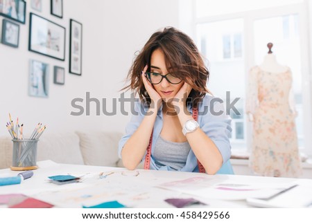 A pretty brunette girl is sitting at the table in the workshop studio. The girl in a blue shirt is busy by looking the sketches in album.