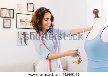 A brunette girl in a blue shirt is working in workshop studio. She is selecting some material for the blue dress.