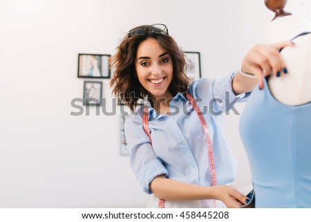 A smiling brunette girl is creating blue dress on the mannequin. She works in a blue shirt. It`s a portrait of a girl in  workshop studio.