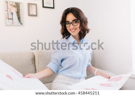 A brunette young girl in a blue shirt is sitting on the sofa in the studio. She has sketches  in her both hands. She is looking to the camera.