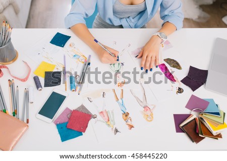 This picture describes the processes of designing clothes. There are hands of a girl drawing sketches on the table. There is creative mess with different stuff on the table.