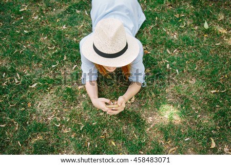 Young pretty girl in straw hat and dress lying on the grass in park  using mobile smart phone top view. Woman typing text message on smart phone using internet