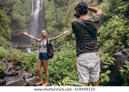Excited young woman standing in front of a waterfall being photographed by her boyfriend. Photographer taking portraits of his girlfriend standing with arms wide open in forest.